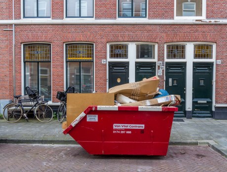 A large red skip bin positioned on a street in front of a row of brick residential buildings in Notting Hill, with the company's contact information displayed on the side. The bin is filled with various waste materials, including flattened cardboard boxes, plastic bags, and what appears to be wooden packaging, all piled to the top with some items draping over the edges. To the left of the bin, two bicycles are secured to a metal railing, and the background features a series of tall, narrow windows with decorative yellow and white window grills. The street pavement in front of the buildings is visibly clean, and the scene suggests a waste collection or clearance activity typical for local rubbish removal services in London neighborhoods like Notting Hill.