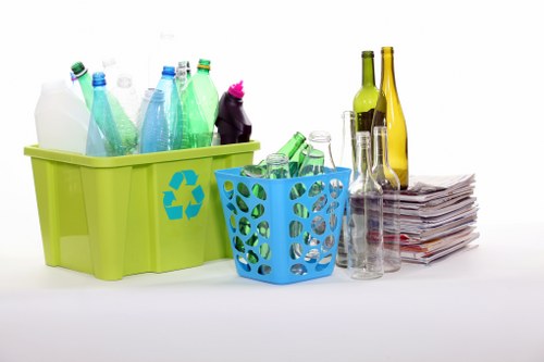 A collection of recyclable waste items arranged on a plain white surface, including a large lime green plastic container with a recycling symbol, holding various plastic bottles in shades of green, blue, and clear with different cap colours, a smaller blue plastic basket filled with additional plastic bottles and containers, and standing next to a neat stack of newspapers or magazines. To the right, there are several empty glass bottles, including green and clear ones, with some grouped together and others slightly separated, against a clean, uncluttered background. The scene suggests an organised approach to waste separation in a residential or commercial setting, likely related to waste or rubbish removal services in Notting Hill, London, highlighting the importance of recycling efforts for local rubbish clearance. The lighting is bright and even, emphasizing the clean and tidy appearance, consistent with professional waste management practices.