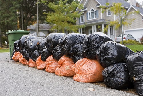 A row of tightly packed black and orange rubbish bags lined up along the edge of a residential street in Notting Hill, with some bags leaning against each other on the pavement. The black bags appear to contain household waste, while the orange bags are likely for recyclable or garden waste. Behind the rubbish bags, there are two green waste or recycling bins positioned near the curb. In the background, a two-storey detached house with light-colored exterior walls, white window frames, and a pitched roof is visible, surrounded by greenery including trees and shrubs. The street is paved with asphalt, and the overall scene suggests a local rubbish removal or clearance operation typical for residential properties in the area, coordinated by a service such as Garden Clearance Notting Hill, which specializes in rubbish disposal and clearance services in the local postcode district.