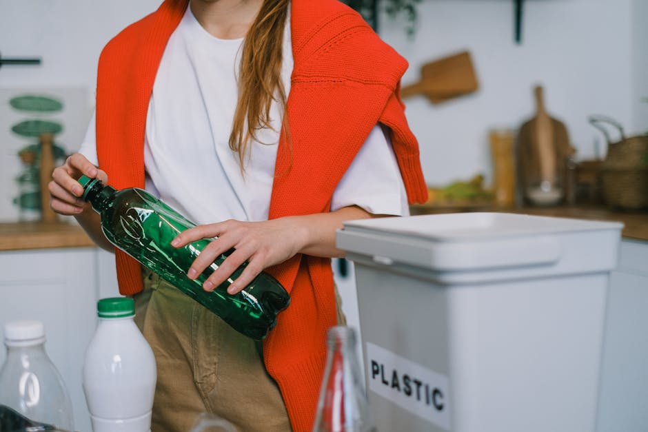 A person with long reddish-brown hair, wearing a white t-shirt and an orange knitted cardigan draped over their shoulders, is holding a green plastic bottle with a black cap in their right hand. The s
