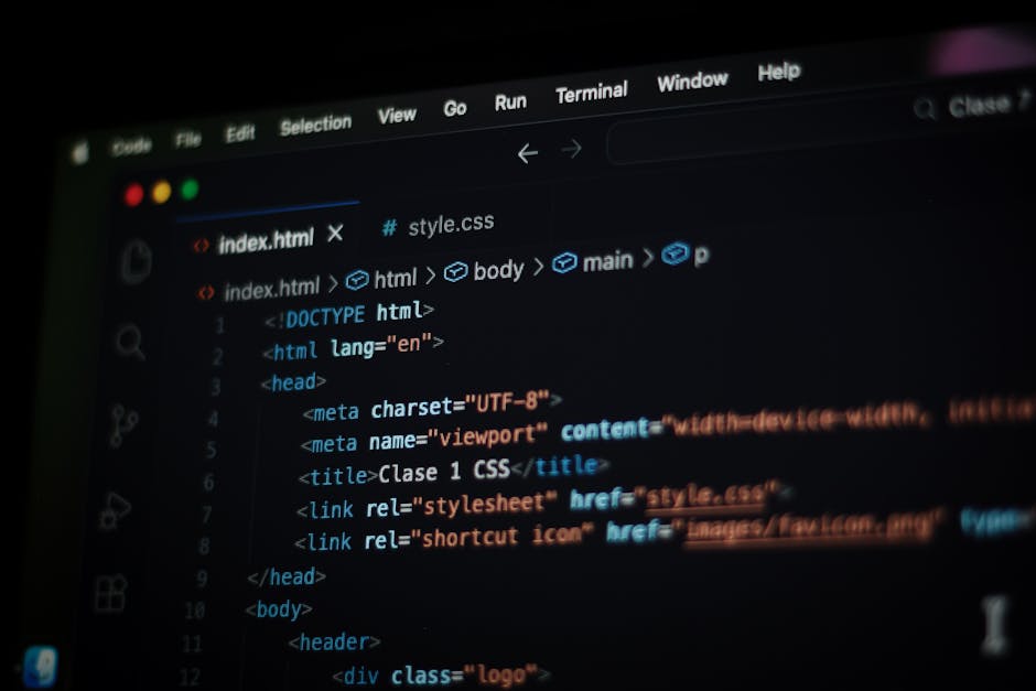 A person seated at a desk working on a laptop with a black screen displaying colorful lines of computer code. The individual, seen from behind, has short dark hair and is wearing a light blue shirt. T