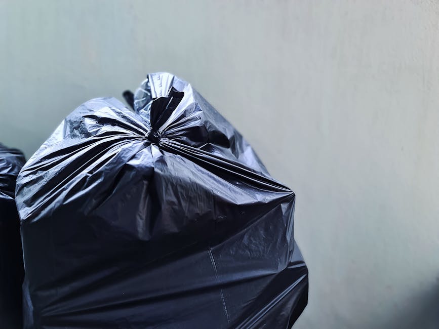A person is seen disposing of brown paper bags filled with trash into a grey rubbish bin with a white lid, positioned on a wooden surface. The person's arm is extended, holding a paper bag above the o